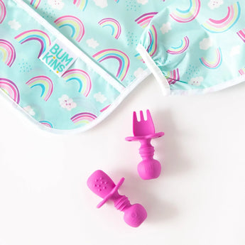 Close-up of dark pink toddler utensils near Bumkins Sleeved Bib: Rainbows on a white background.