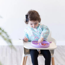 Load image into Gallery viewer, A toddler wears a Bumkins Sleeved Bib: Rainbows, eating fruit with a spoon from a purple plate in a high chair.