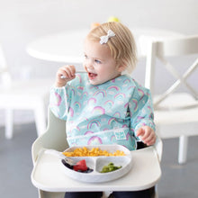 Load image into Gallery viewer, A toddler in a high chair, wearing a Bumkins Sleeved Bib: Rainbows, happily enjoys pasta, strawberries, and broccoli from a gray plate.