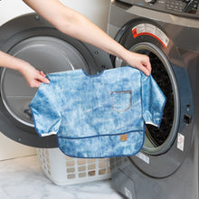 Load image into Gallery viewer, A person holds Bumkins Sleeved Bib: Vintage Denim in front of an open front-loading washing machine.