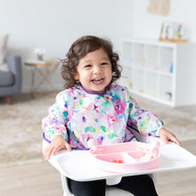 Load image into Gallery viewer, A curly-haired girl smiles in a high chair, wearing a floral bib and eating from a Bumkins Silicone Grip Dish: Pink in a bright playroom.