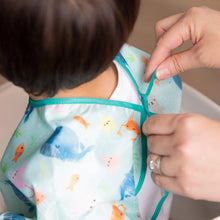Load image into Gallery viewer, Close-up of hands adjusting the ribbon closure of a Bumkins Sleeved Bib: Ocean Life around a child's neck.