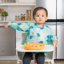 Load image into Gallery viewer, A toddler in a Bumkins Sleeved Bib: Ocean Life sits in a high chair with an orange divided plate in a kitchen.