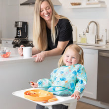 Load image into Gallery viewer, A smiling woman watches a happy toddler in a Bumkins Sleeved Bib: Ocean Life in a high chair, with an orange plate and utensils on the tray.