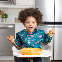 Load image into Gallery viewer, A curly-haired toddler sits in a high chair, wearing a Bumkins Sleeved Bib: Jungle, holding a spoon by an orange divided plate.