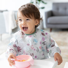 Load image into Gallery viewer, A girl wearing a floral bib in a high chair smiles as she self feeds from Bumkins Silicone First Feeding Set: Pink in a cozy living room.