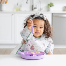 Load image into Gallery viewer, A toddler excitedly holds a spoon while wearing a Bumkins Sleeved Bib: Floral, sitting at a white table with a divided purple plate.
