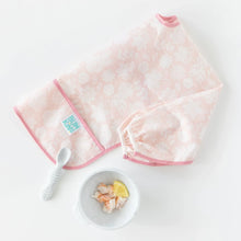 Load image into Gallery viewer, Bumkins Sleeved Bib: Lace lays folded next to a white spoon and a white bowl with shrimp and lemon on a white background.