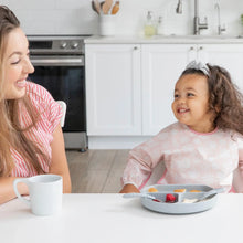 Load image into Gallery viewer, A woman and girl wearing a Bumkins Sleeved Bib: Lace smile at each other at a white table in a bright kitchen; a divided plate and cup nearby.