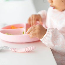 Load image into Gallery viewer, Close-up of a toddler in a Bumkins Sleeved Bib: Lace eating from a pink divided plate at a white table.