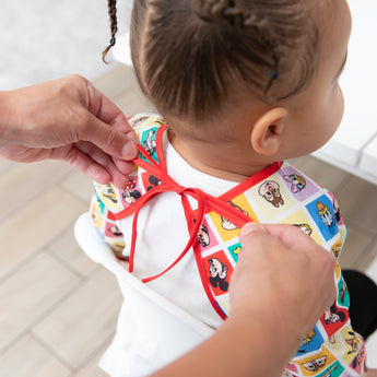 An adult ties the neck closure of Bumkins Sleeved Bib: Mickey & Friends Check Mates on a child with braided hair.
