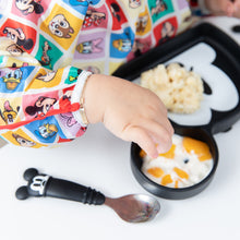 Load image into Gallery viewer, A child in Bumkins Sleeved Bib: Mickey & Friends Check Mates enjoys baby-led weaning from a Mickey-shaped plate.
