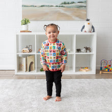 Load image into Gallery viewer, A toddler in a Bumkins Sleeved Bib: Mickey & Friends Check Mates smiles before a toy-filled shelf in a cozy room.