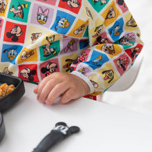 Load image into Gallery viewer, A child in Bumkins Sleeved Bib: Mickey & Friends Check Mates sits at a white table with a black dish and utensil.