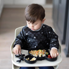 Load image into Gallery viewer, A toddler in a high chair in a Bumkins Sleeved Bib: Mickey Mouse Icon Black + White eats from a divided plate filled with rice and fruit.