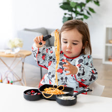 Load image into Gallery viewer, A toddler wearing a Bumkins Sleeved Bib: Mickey Mouse eats spaghetti with a fork from a Mickey Mouse-shaped plate at a white table.