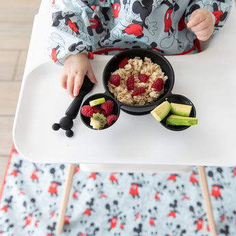 A baby in a high chair wearing a blue bib eats oatmeal & fruit from Bumkins Silicone First Feeding Set: Mickey Mouse Classic Black.