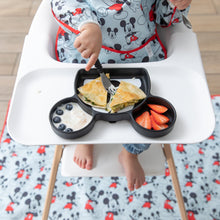Load image into Gallery viewer, A child in a Bumkins Sleeved Bib: Mickey Mouse sits in a high chair with a matching mat on the floor, eating from a Mickey-shaped plate.