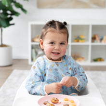 Load image into Gallery viewer, A child with pigtails smiles in a Bumkins Sleeved Bib: Mickey + Minnie Sunny Side Up in a high chair with snacks on a pink plate.