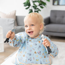 Load image into Gallery viewer, A blonde toddler with a fork and spoon sits in a bright room, wearing Bumkins Sleeved Bib: Mickey + Minnie Sunny Side Up.