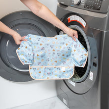 Load image into Gallery viewer, A person holds a Bumkins Sleeved Bib: Mickey + Minnie Sunny Side Up in front of a front-load washer.