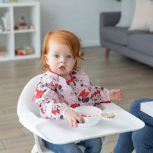 Load image into Gallery viewer, A young girl wearing a Bumkins Sleeved Bib: Minnie Mouse in a highchair eating from the Silicone First Feeding Set: Vanilla Sprinkle.
