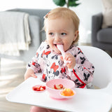 A young girl wearing a Bumkins Sleeved Bib: Minnie Mouse in a highchair eating from the Silicone First Feeding Set: Minnie Mouse Light Pink.
