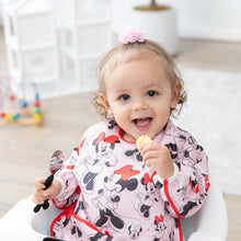 Load image into Gallery viewer, A young girl smiles, wearing a Bumkins Sleeved Bib: Minnie Mouse in a high chair while holding a spoon and a snack.