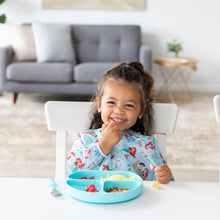Load image into Gallery viewer, A child wearing Bumkins Sleeved Bib: Ariel smiles while eating from a blue sectioned plate at a white table; a gray couch in the background.