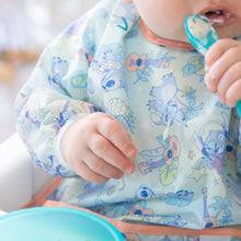 Load image into Gallery viewer, A baby enjoys self-feeding with a spoon, wearing a Bumkins Sleeved Bib: Aloha Stitch made of waterproof fabric.