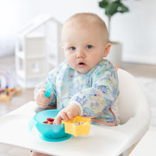 Load image into Gallery viewer, A baby in a high chair, focused on baby-led weaning, wears a charming Bumkins Sleeved Bib: Aloha Stitch against a bright backdrop.