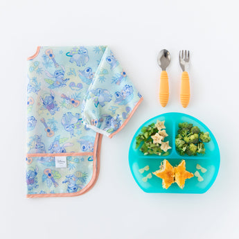 Bumkins Sleeved Bib: Aloha Stitch lays folded next to a blue divided plate with food and orange utensils on a white background.