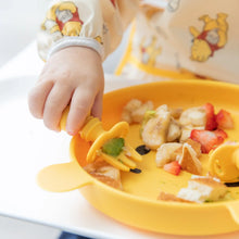 Load image into Gallery viewer, Close-up of a child wearing a Bumkins Sleeved Bib: Winnie Loves Hunny, their hand using a fork to eat diced fruits from a yellow plate.