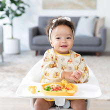Load image into Gallery viewer, A toddler in a high chair claps hands, wearing a Bumkins Sleeved Bib: Winnie Loves Hunny, with food on a plate in a bright room.