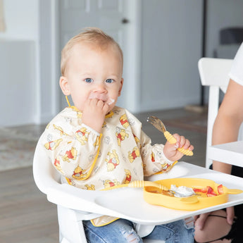 A baby in a high chair, wearing a Bumkins Sleeved Bib: Winnie Loves Hunny, enjoys baby-led weaning with a yellow fork and plate.