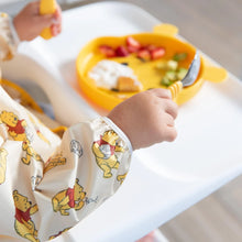 Load image into Gallery viewer, Close-up of a child in a high chair wearing a Bumkins Sleeved Bib: Winnie Loves Hunny, holding a fork and spoon over a yellow plate.