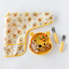 Load image into Gallery viewer, Bumkins Sleeved Bib: Winnie Loves Hunny lays folded next to a yellow bear-shaped plate of food and yellow utensils on a white background.