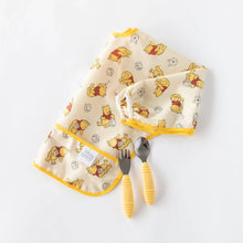 Load image into Gallery viewer, Bumkins Sleeved Bib: Winnie Loves Hunny lays folded on a white background with a yellow fork and spoon.