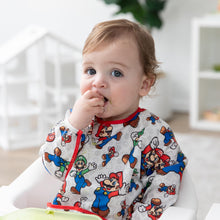 Load image into Gallery viewer, A light-haired boy wearing a Bumkins Sleeved Bib: Super Mario™ Classic in a high chair places food in their mouth.