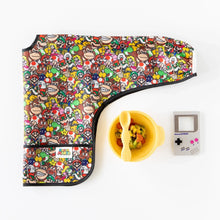 Load image into Gallery viewer, Bumkins Sleeved Bib: Super Mario™ Mashup lays folded by a yellow bowl with spoon, and Nintendo Game Boy teether on a white background.
