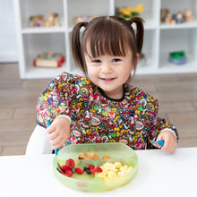Load image into Gallery viewer, A smiling girl wearing a Bumkins Sleeved Bib: Super Mario™ Mashup eats breakfast from a green plate in front of toy-filled shelves.