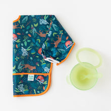 Load image into Gallery viewer, Bumkins Sleeved Bib: Jungle lays folded next to a green transparent bowl & spoon set on a white background.