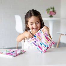 Load image into Gallery viewer, A girl in a white shirt smiles, holding a Bumkins Reusable Snack Bag, Large 2-Pack: Watercolor & Brush Strokes as the other sits on a table.
