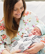 A woman beams holding her sleeping newborn in a Modern Burlap Organic Swaddle, Cactus, wearing a floral robe made of GOTS certified cotton.