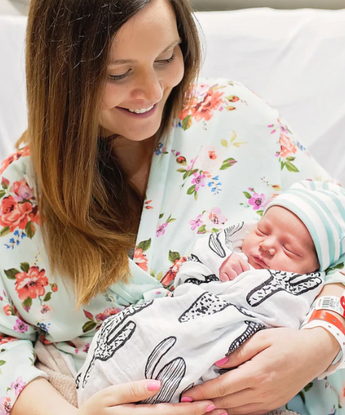 A woman beams holding her sleeping newborn in a Modern Burlap Organic Swaddle, Cactus, wearing a floral robe made of GOTS certified cotton.