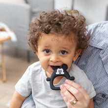Load image into Gallery viewer, A curly-haired toddler chews a Bumkins Silicone Flat Teether: Mickey Mouse, while sitting on an adult's lap in a cozy room.