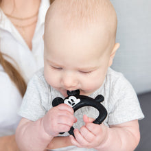 Load image into Gallery viewer, A bald baby in a gray shirt sits on a lap, eagerly chewing on a Bumkins Silicone Flat Teether: Mickey Mouse.