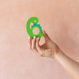 A hand holds Bumkins Silicone Teether: Dinosaur in front of a textured light orange wall.