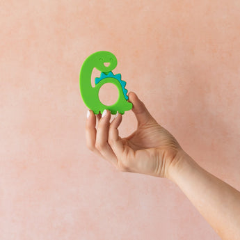 A hand holds Bumkins Silicone Teether: Dinosaur in front of a textured light orange wall.
