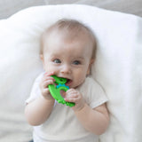 A baby chews on a Bumkins Silicone Teether: Dinosaur, lying on a white blanket.